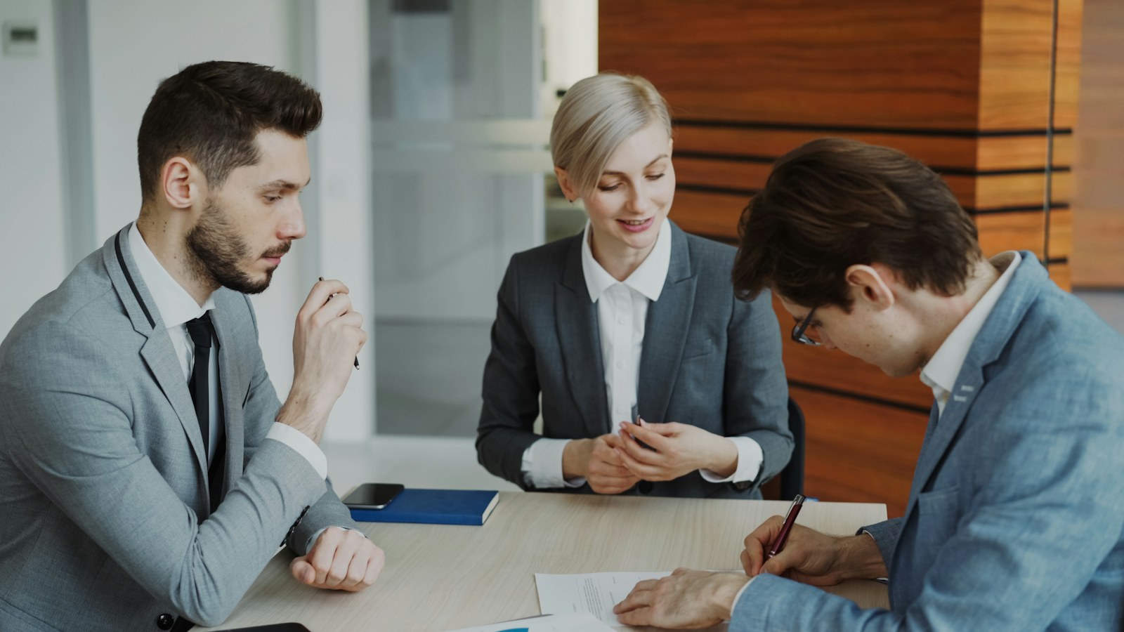 A client signs a contract at an office table while two agency staff look on.