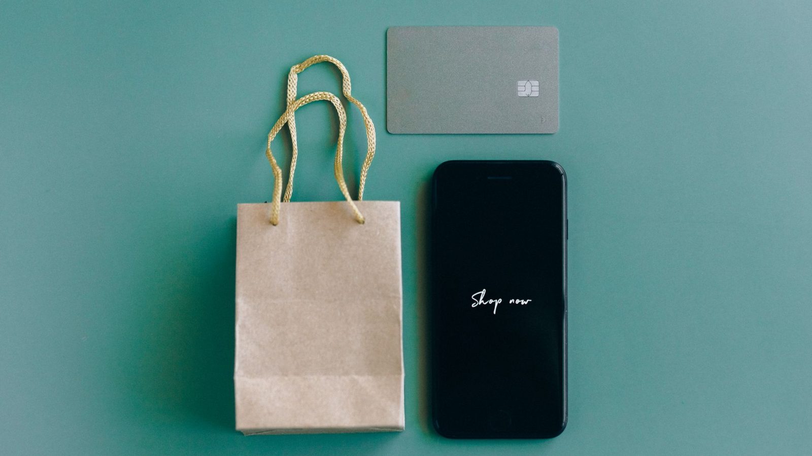 A smartphone showing the words "Shop now" beside a paper shopping bag and a payment card on a teal background, standing for online shopping.